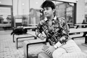attraktiv och stilig snygg indisk man bär skjorta, håller mobiltelefon och sitter på terrassen på moderna loungecafé på sin arbetsrast. foto