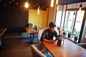 självsäker ung indisk man i svart skjorta sitter på café och läsa menyn. foto