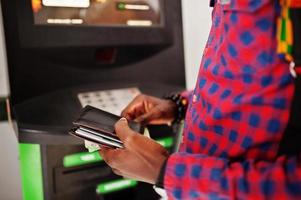 afrikansk amerikansk man i rutig skjorta med ryggsäck som håller plånboken i handen mot bankomat. svart man resenär. foto
