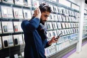 indisk skäggman kundköpare i mobiltelefonbutik lyssna på musik från trådlösa hörlurar anslut till sin smartphone. sydasiatiska folk och teknik koncept. mobiltelefon butik. foto