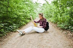 porträtt av en positiv ung vacker blondin som sitter på marken med en karta i händerna i skogen. foto