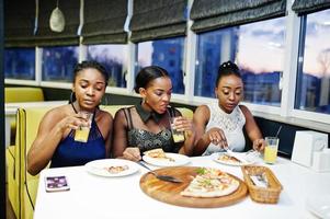 tre afrikansk kvinna i klänning poserar på restaurang, äter pizza och dricker juice. foto