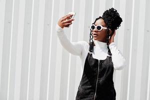afrikansk kvinna i jeansoverallkjol, vita solglasögon poserade mot en vit stålvägg och gör selfie via telefon. foto