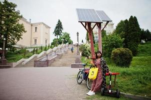 vacker afrikansk amerikansk kvinna som laddar segway eller hoverboard av solpaneler. svart flicka med gul duk eco väskor återvinning symbol. foto