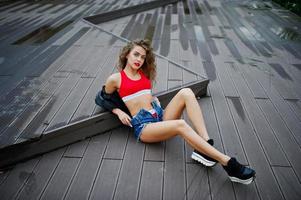 sexig lockig modelltjej i röd topp, jeansshorts, skinnjacka och sneakers poserade på golvet. foto