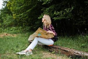 porträtt av en positiv ung vacker blondin som sitter på marken med en karta i händerna i skogen. foto