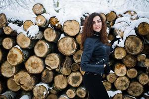 elegans lockig tjej i skinnjacka på snöig skogspark på vintern mot stubbar. foto