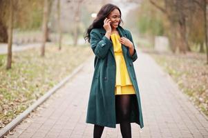 snygg afrikansk amerikansk kvinna i grön kappa och gul klänning poserade mot höstparken och pratar i telefon. foto