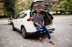 framgångsrik arab man bär i randig skjorta och solglasögon sitter i bagageutrymmet på sin vita suv bil med mobiltelefon. foto