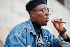 närbild porträtt av afrikansk amerikansk man i jeansjacka, basker och glasögon, röka cigarr. foto