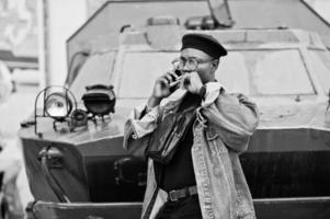 afrikansk amerikansk man i jeansjacka, basker och glasögon, med cigarr poserad mot btr militära pansarfordon, och talar i mobiltelefon. foto