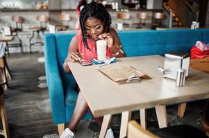 attraktiv afrikansk amerikansk kvinna sitter vid bordet på café med latte och tittar på mobiltelefon. foto