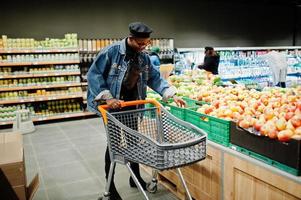 snygg casual afrikansk amerikansk man vid jeansjacka och svart basker på den ekologiska delen av snabbköpet med kundvagnen. foto