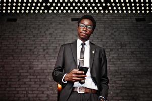 affärsafrikansk amerikansk man bär på svart kostym och glasögon på kontoret tittar på mobiltelefon mot futuristiska ljus. foto