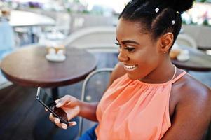 afrikansk amerikansk flicka sitter på bordet av caffe med mobiltelefon. foto