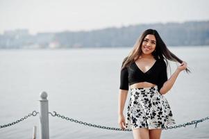 söt latino modell tjej från ecuador bär på svarta toppar och kjol poserade mot sjön. foto