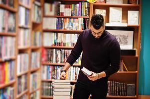 lång smart arabisk student man, bära på violett turtleneck och glasögon, på biblioteket med trave böcker. foto