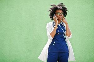 porträtt av afrikansk amerikansk kvinnlig läkare med stetoskop bär labbrock, talar med mobiltelefon. foto