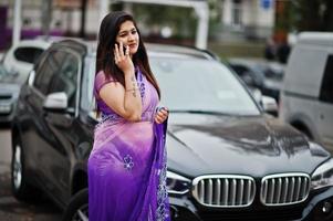 indisk hinduisk flicka på traditionella violett saree poserade på gatan mot svart business suv bil och talar i mobiltelefon. foto
