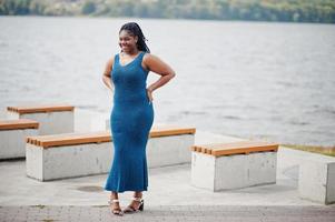 afroamerikansk mörkhyad plus size-modell poserad i en blå glänsande klänning mot havet. foto