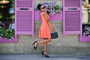 afrikansk amerikansk tjej i solglasögon, persika klänning och handväska poserade mot lila fönster. foto