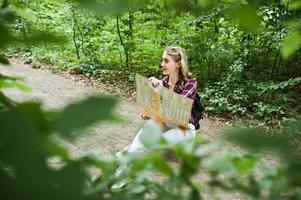 porträtt av en positiv ung vacker blondin som sitter på marken med en karta i händerna i skogen. foto