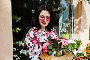 sommar porträtt av brunett flicka i rosa glasögon och hatt mot blommor butik. foto