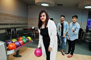 sydasiatisk kvinna står vid bowlinghall med boll på händerna. flickan förbereder sig för ett kast. vänner stöttar henne högt. foto