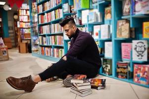 lång smart arab student man, bära på violett turtleneck och glasögon, på biblioteket sitter mot bokhyllor. foto