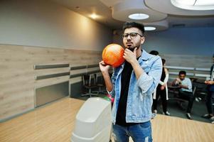 snygg asiatisk man i jeansjacka och glasögon stående vid bowlinghall med boll till hands. foto