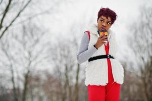 afrikansk amerikansk kvinna i röda byxor och vit pälsjacka poserade på vinterdagen mot snöig bakgrund och håller en kopp kaffe. foto