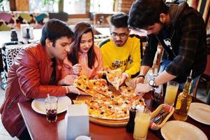 grupp asiatiska vänner som äter pizza under fest på pizzeria. glada indianer har roligt tillsammans, äter italiensk mat och sitter på soffan. foto