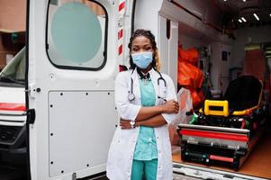 afrikansk amerikansk kvinnlig sjukvårdare i ansiktsskyddande medicinsk mask som står framför ambulansbil. foto
