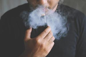 foto porträtt ung man med skägg hålla och röka sin elektroniska cigarett blåser en ström av rök sprids. mörk svart, grå bakgrund