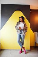 fashionabla afrikansk amerikansk kvinna i svart mössa chillande i mysigt café och päls mot gul vägg. foto