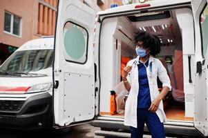 afrikansk amerikansk kvinnlig sjukvårdare i ansiktsskyddande medicinsk mask som står framför ambulansbil. foto