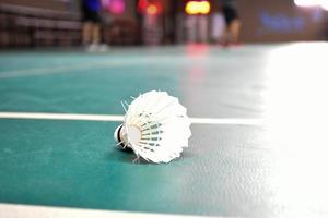 gräddvit badmintonfjärdeboll och racket med neonljusskuggning på grönt golv i inomhusbadmintonbana, suddig badmintonbakgrund, kopieringsutrymme. foto