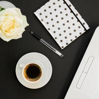 platt låg, ovanifrån kontorsbord skrivbord. arbetsyta med laptop, vit ros, prickiga dagbok och kaffemugg på svart bakgrund. foto