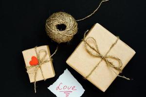 handgjord presentförpackning insvept i hantverkspapper med rött trähjärta, rep och rosett på svart bakgrund. ovanifrån, platt låg foto