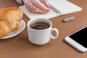 kaffepaus med croissant och espress. kvinna skriver på anteckningsboken. frilansare på jobbet foto