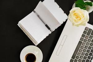 platt låg, ovanifrån kontorsbord skrivbord. arbetsyta med laptop, vit ros, öppen dagbok och kaffemugg på svart bakgrund. foto