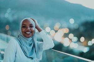 afrikansk muslim kvinna i de natt på en balkong leende på de kamera med stad bokeh lampor i de bakgrund. foto