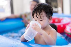 porträtt bebis 1-2 år gammal. asiatisk barn pojke dricka vatten med plast glas. förtjusande unge stående i sudd slå samman. i sommar tid. barn dryck vatten till släcka hans törst och Häftigt av värme. foto