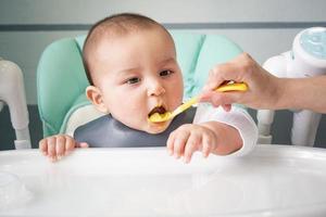 mamma matar de bebis med en sked av vegetabiliska puré på de barns matning tabell. bebis aptit, friska näring, introduktion av komplementär livsmedel. copyspace, falsk upp foto