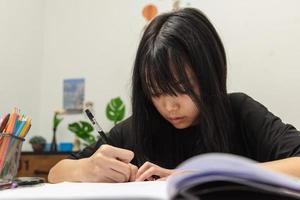 asiatisk studentflicka skriver läxor och läser bok vid skrivbordet foto