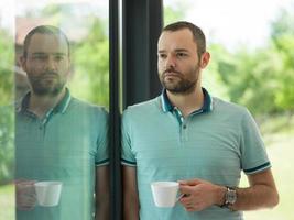 ung man dricka morgon- kaffe förbi de fönster foto