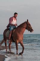 en modern man i sommar kläder åtnjuter ridning en häst på en skön sandig strand på solnedgång. selektiv fokus foto
