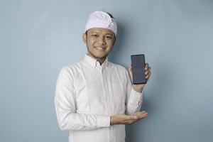 upphetsad balinesisk man bär traditionell pannband eller udeng och vit skjorta pekande på de kopia Plats på hans smartphone, isolerat förbi blå bakgrund foto