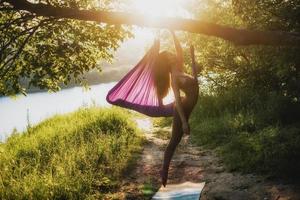 en ung kvinna gymnast är engagerad i antenn yoga, använder sig av en kombination av traditionell yoga poserar, pilates och dansa använder sig av en hängmatta på solnedgång i natur. friska livsstil. foto