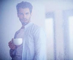 avslappnad ung man dryck först morgon- kaffe med regn droppar på fönster foto